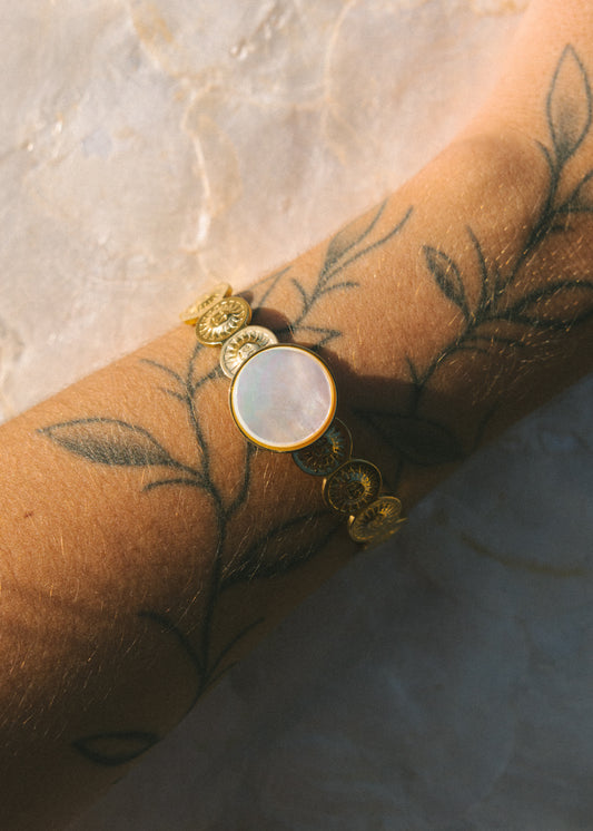 Gold bracelet with a circular gemstone on a wrist with tattoos, against a marble background