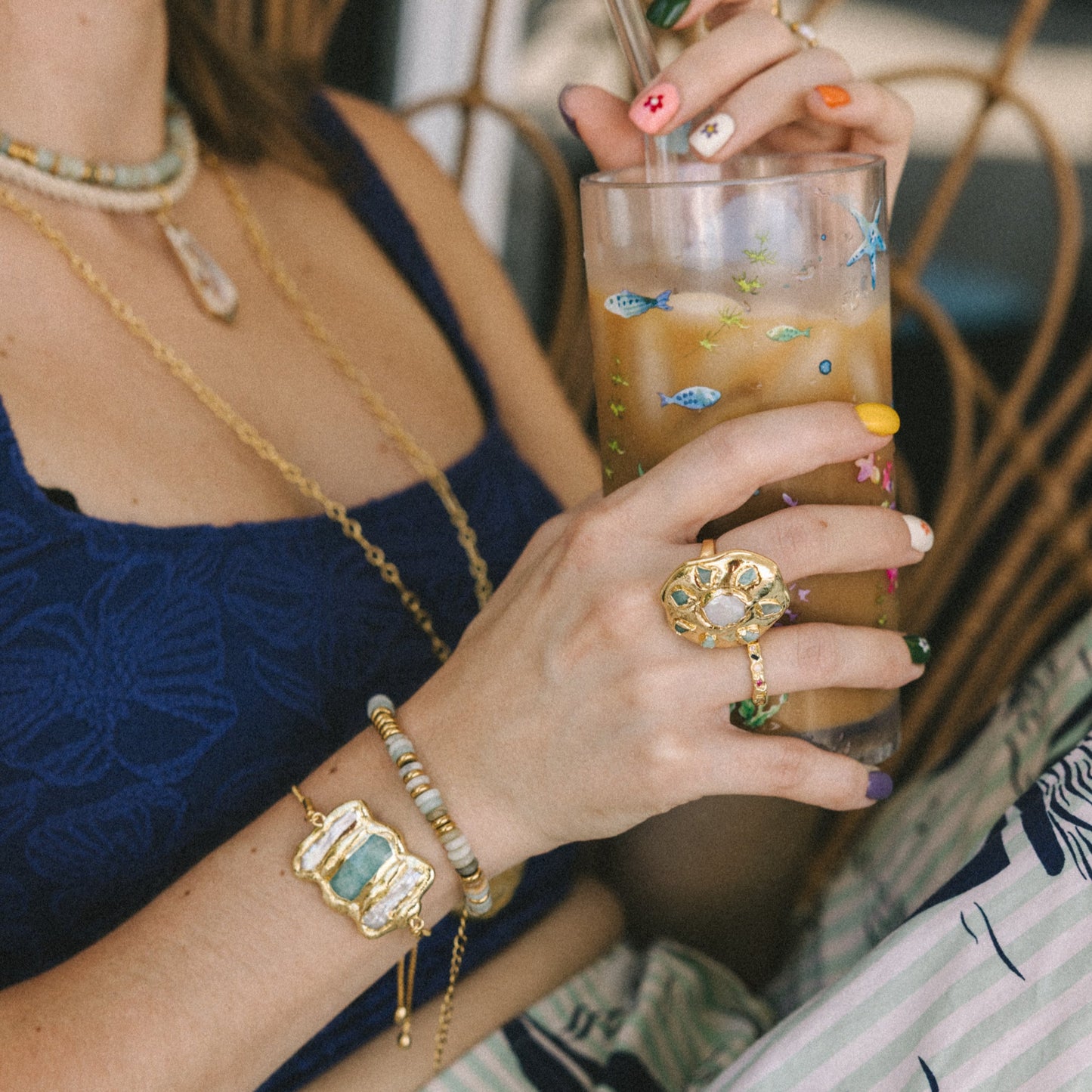 Person holding a glass of iced drink with colorful ice cubes, wearing gold jewelry.