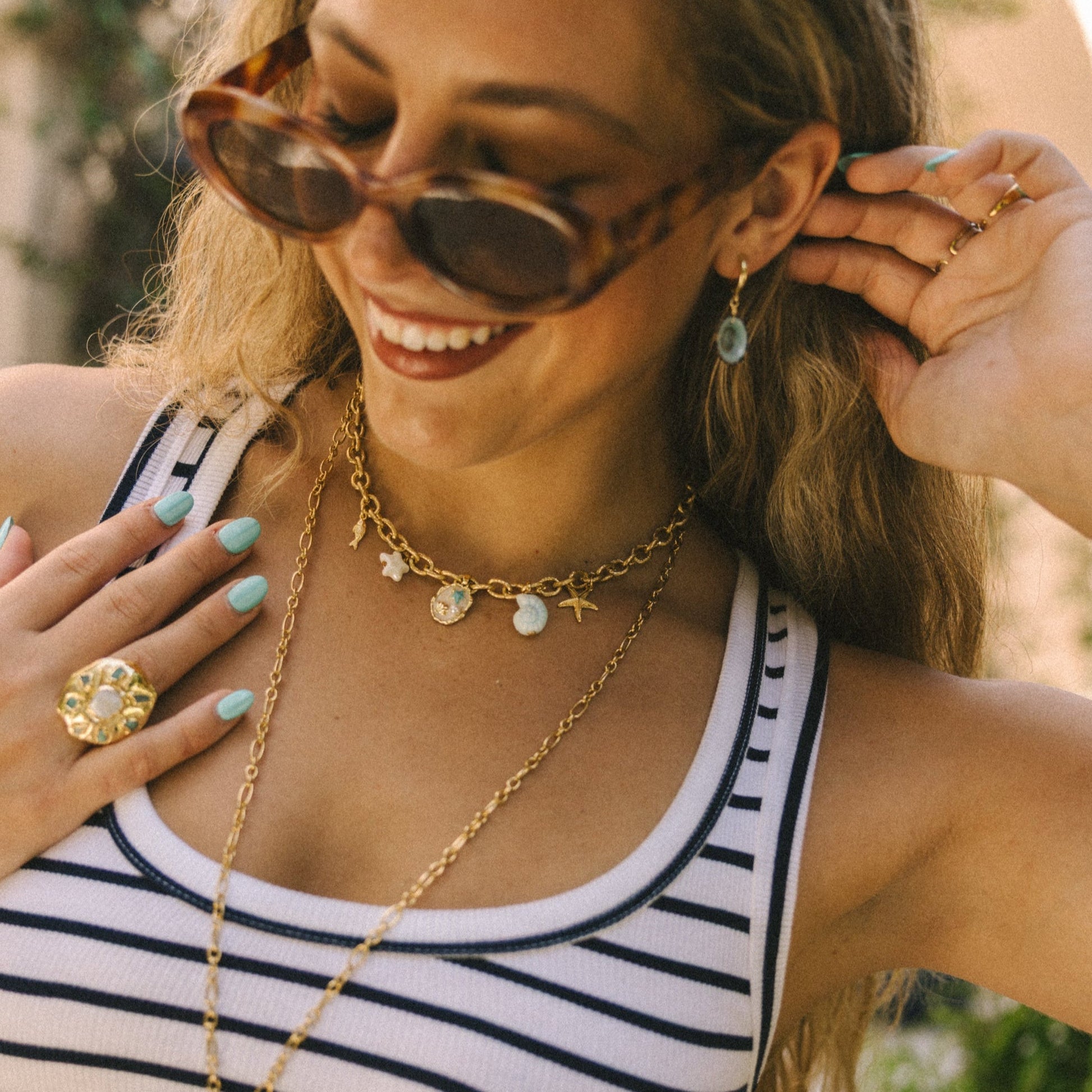 Woman wearing sunglasses, a striped tank top, and layered necklaces with a blurred outdoor background