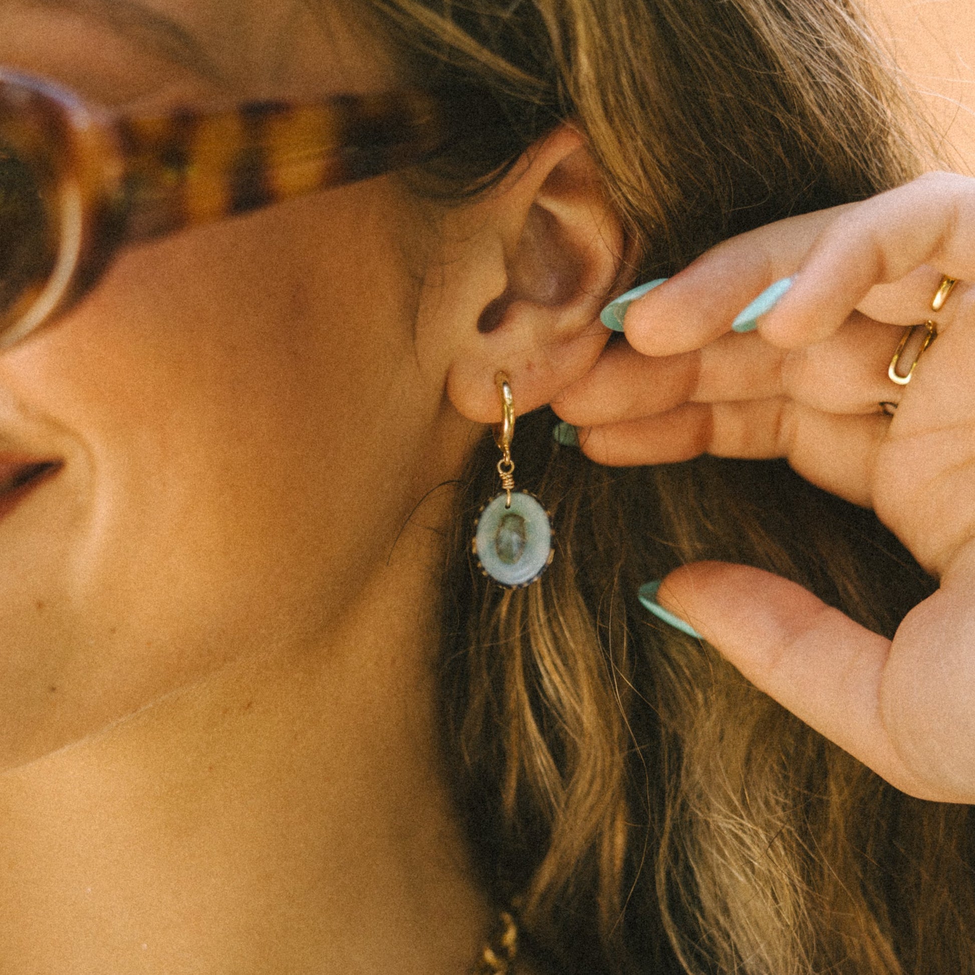 Close-up of a person wearing gold earrings with green gemstones, holding a small green object near their ear.