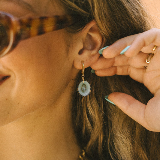 Close-up of a person wearing gold earrings with green gemstones, holding a small green object near their ear.