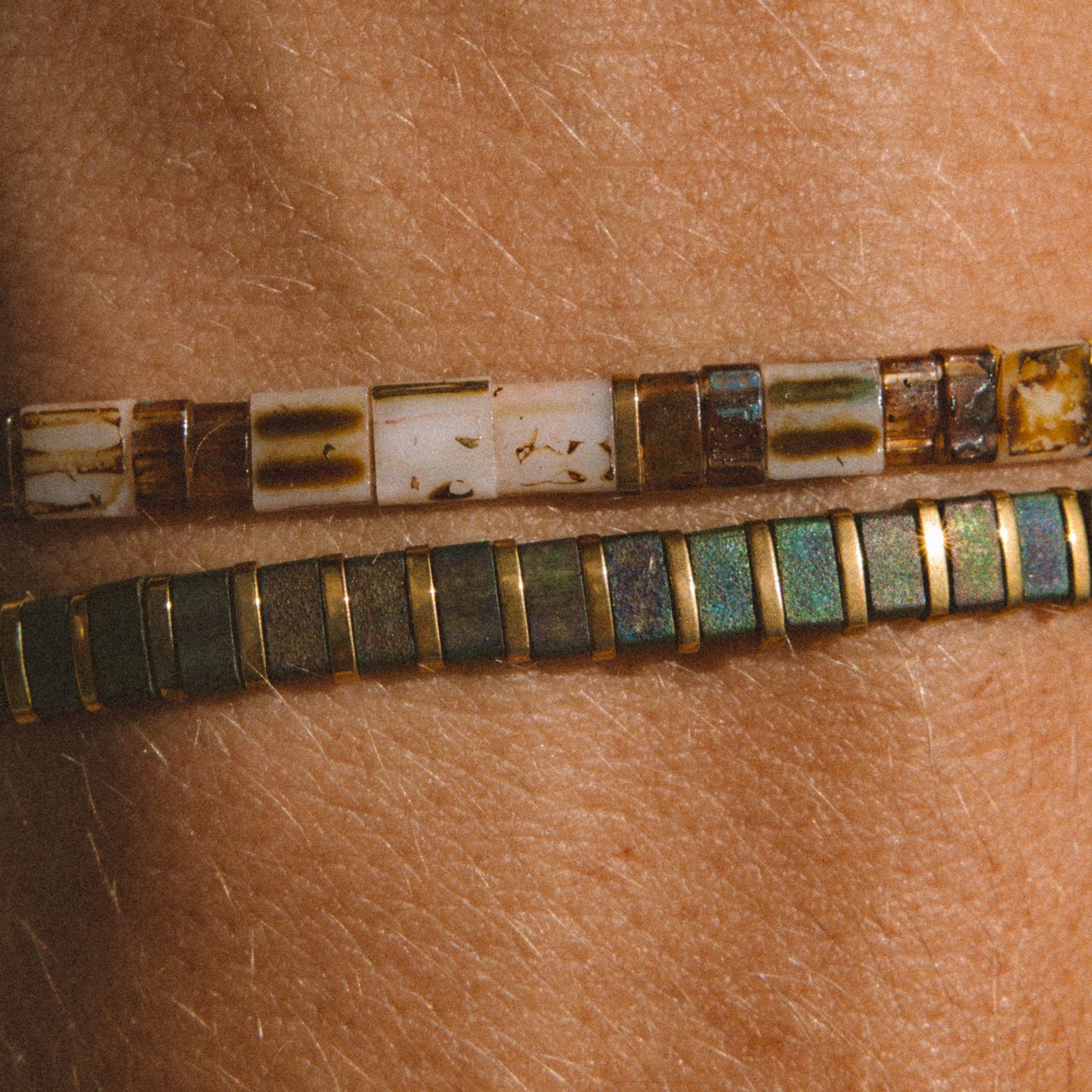 Two beaded bracelets worn on a person's wrist, featuring rectangular beads in earthy brown, cream, and green with gold-toned borders.