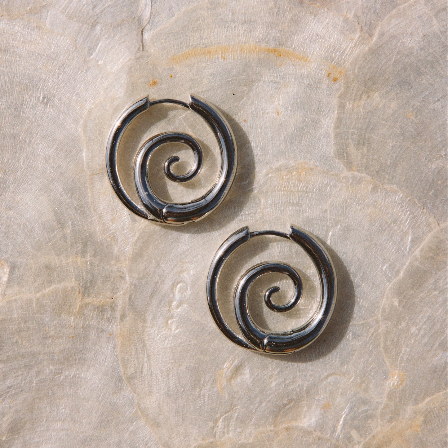A pair of dark metallic spiral hoop earrings resting on a textured beige stone surface.