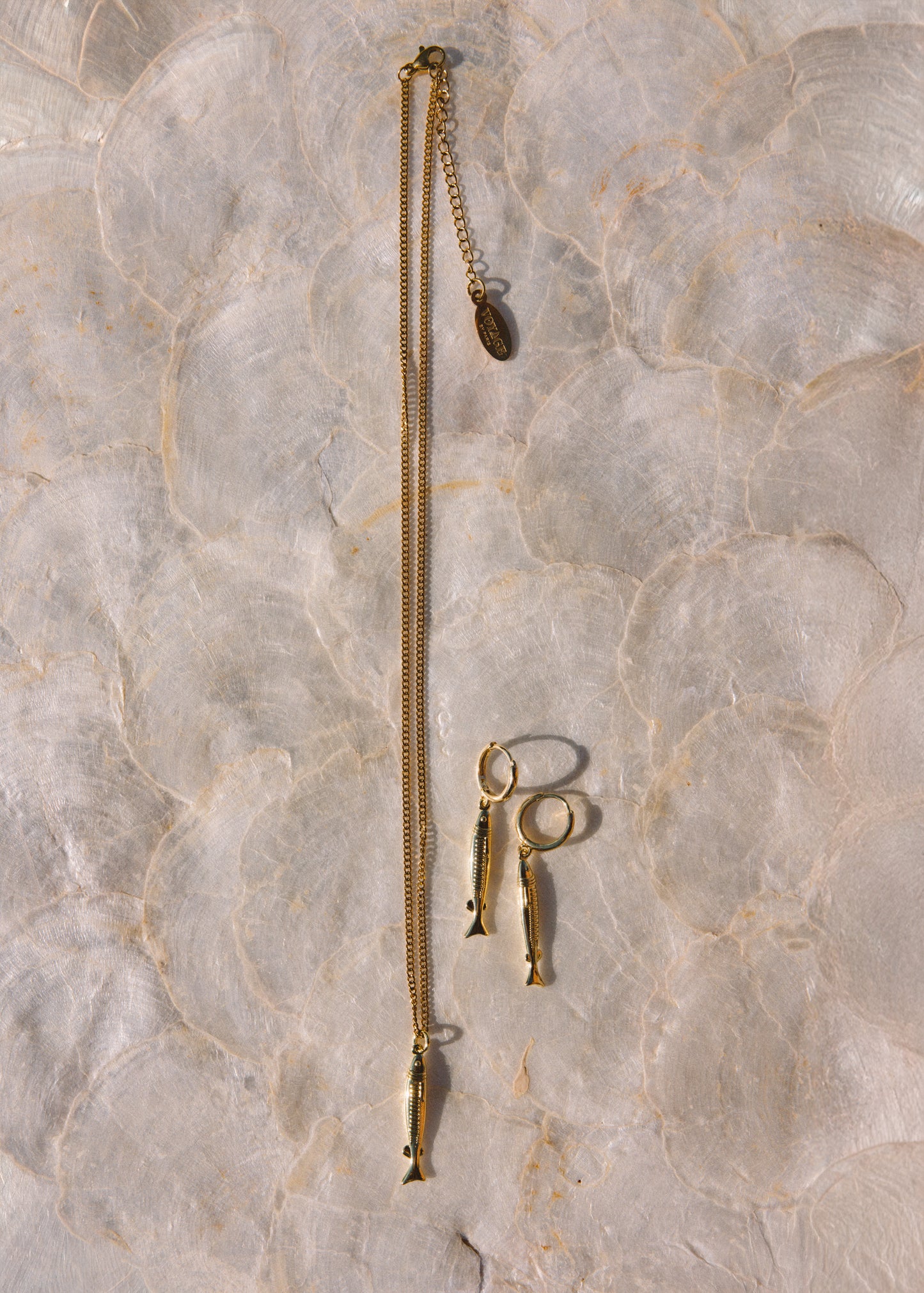 A gold-toned jewelry set featuring a long chain necklace with a small fish-shaped pendant, matching fish-shaped drop earrings, and a small ring, laid flat on a light beige marble surface.