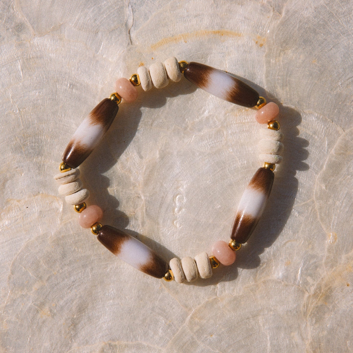 A beaded stretch bracelet featuring alternating white and brown marbled beads, small pale pink beads, and gold spacer beads, displayed on a textured beige stone surface.