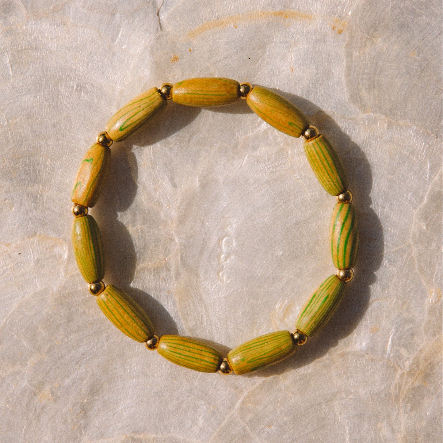A beaded stretch bracelet made of olive green and yellow-green seed-like beads separated by small gold spacer beads, displayed on a light beige stone surface.