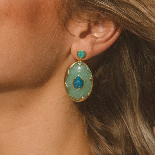 A close-up of a model's ear wearing a gold-toned drop earring with a light green oval stone and a central blue stone accent.