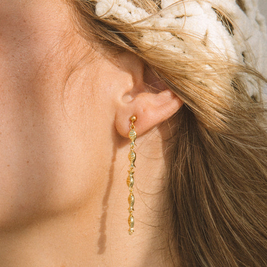 A close-up of a woman's ear wearing a delicate gold dangle earring with small faceted beads, against warm sunlight and a white knit headband.