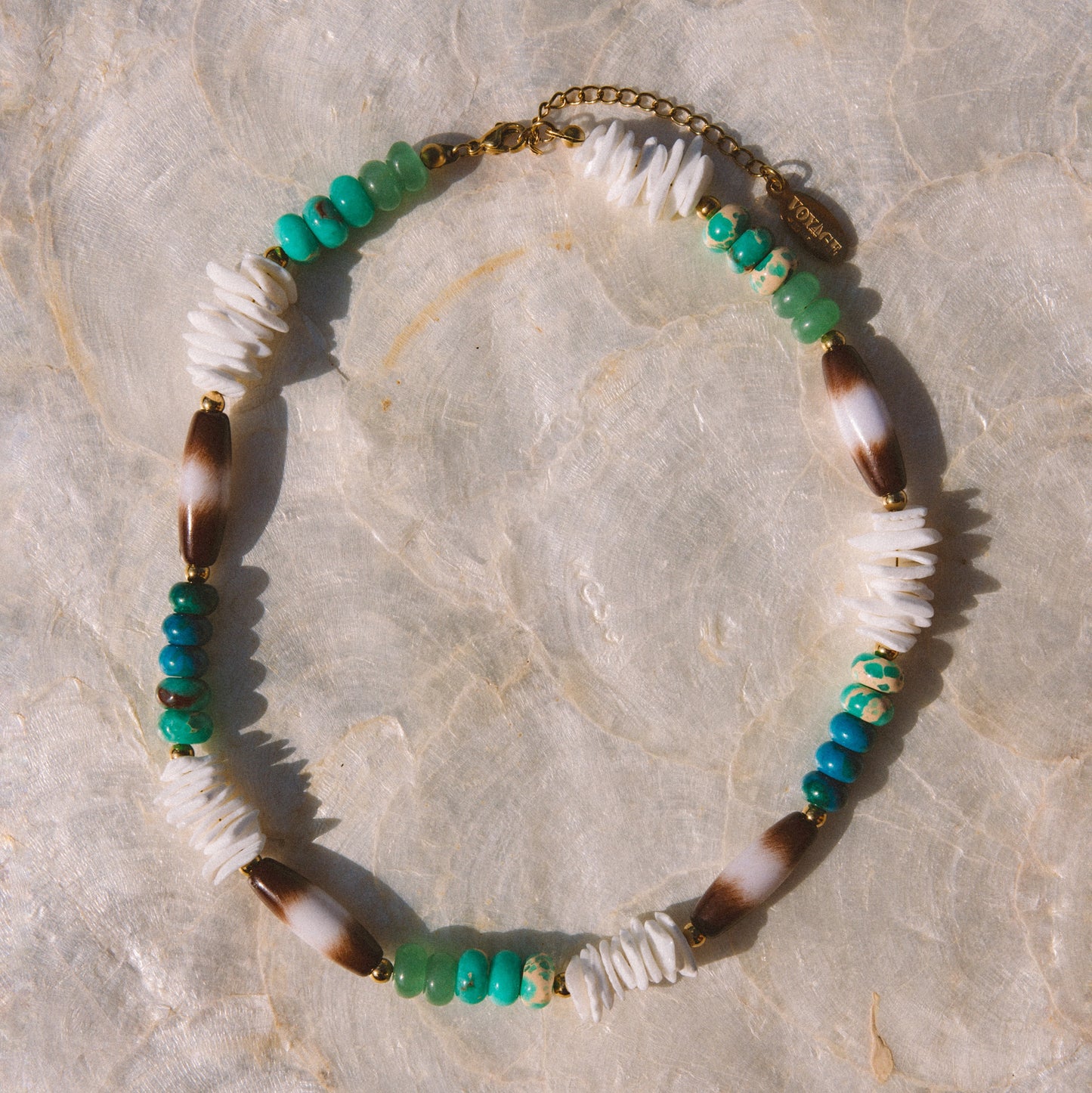 A bohemian-style beaded necklace featuring white beads, green and turquoise beads, brown and white marbled beads, and gold-toned metal accents, displayed on a light beige stone surface.