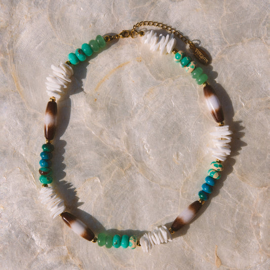 A bohemian-style beaded necklace featuring white beads, green and turquoise beads, brown and white marbled beads, and gold-toned metal accents, displayed on a light beige stone surface.
