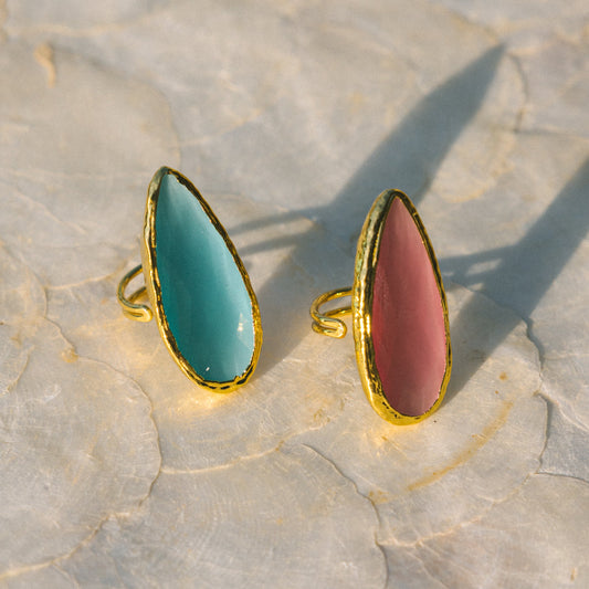 Two gold-rimmed rings with turquoise and pink stones on a textured surface
