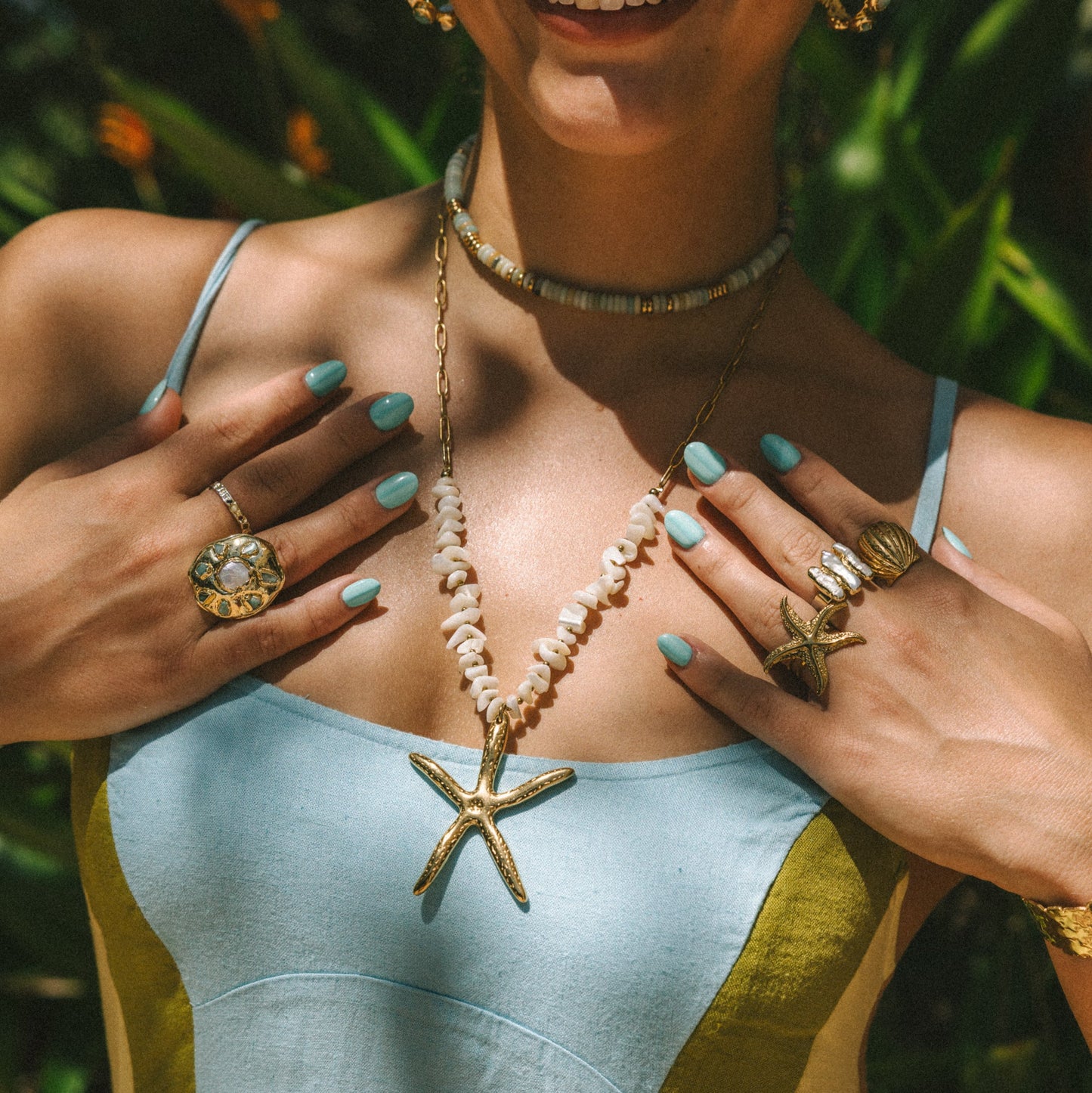 Woman wearing a light blue top with a starfish pendant, surrounded by green foliage.