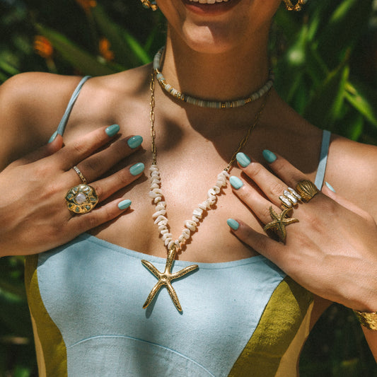 Woman wearing a light blue top with a starfish pendant, surrounded by green foliage.