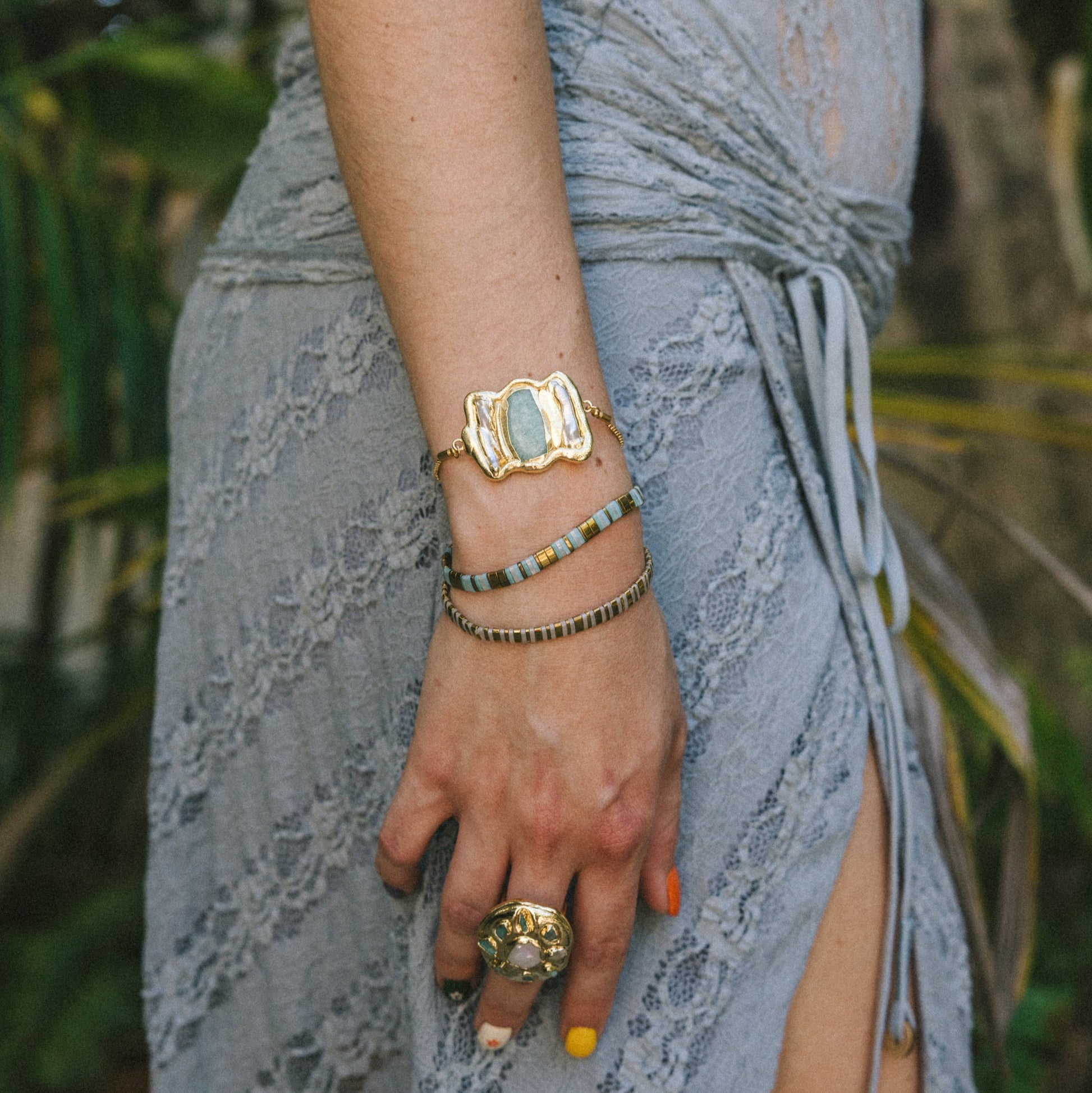 Person wearing a gold bracelet and ring with a blurred natural background