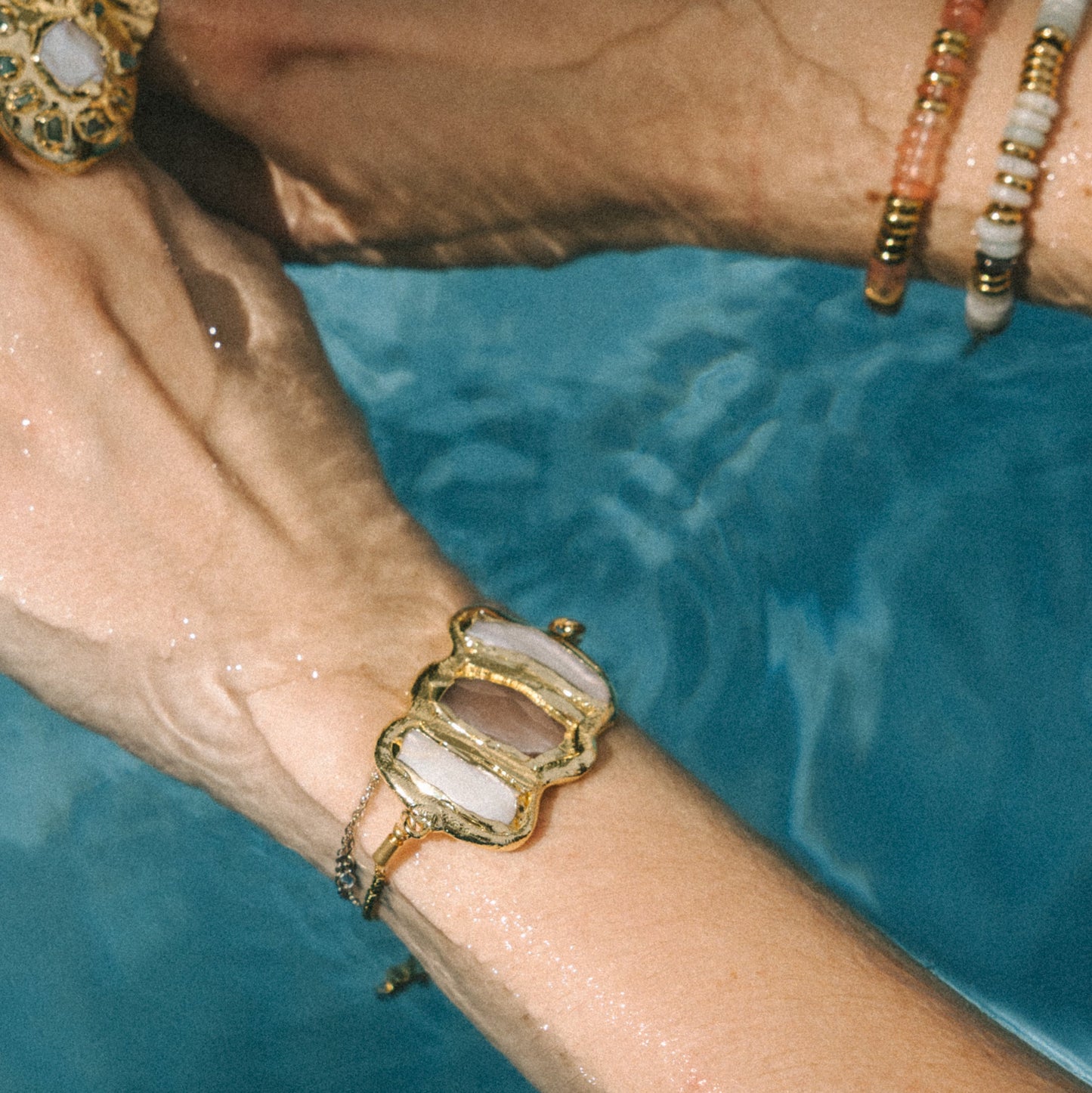 Close-up of a wrist wearing a gold bracelet with a natural stone, against a blue water background.