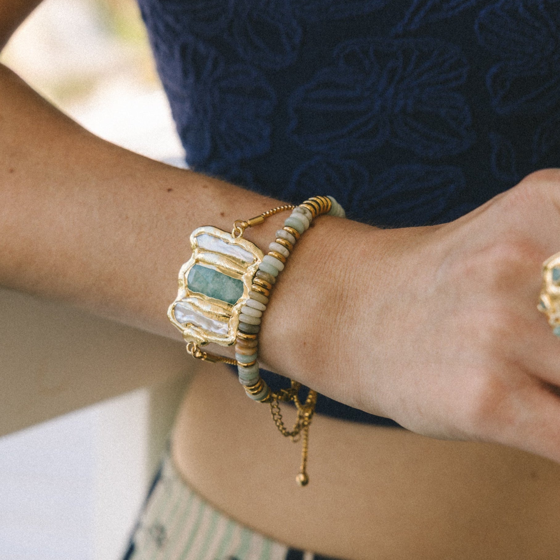 Close-up of a person's arm with gold bracelets and rings, wearing a blue top and patterned pants.