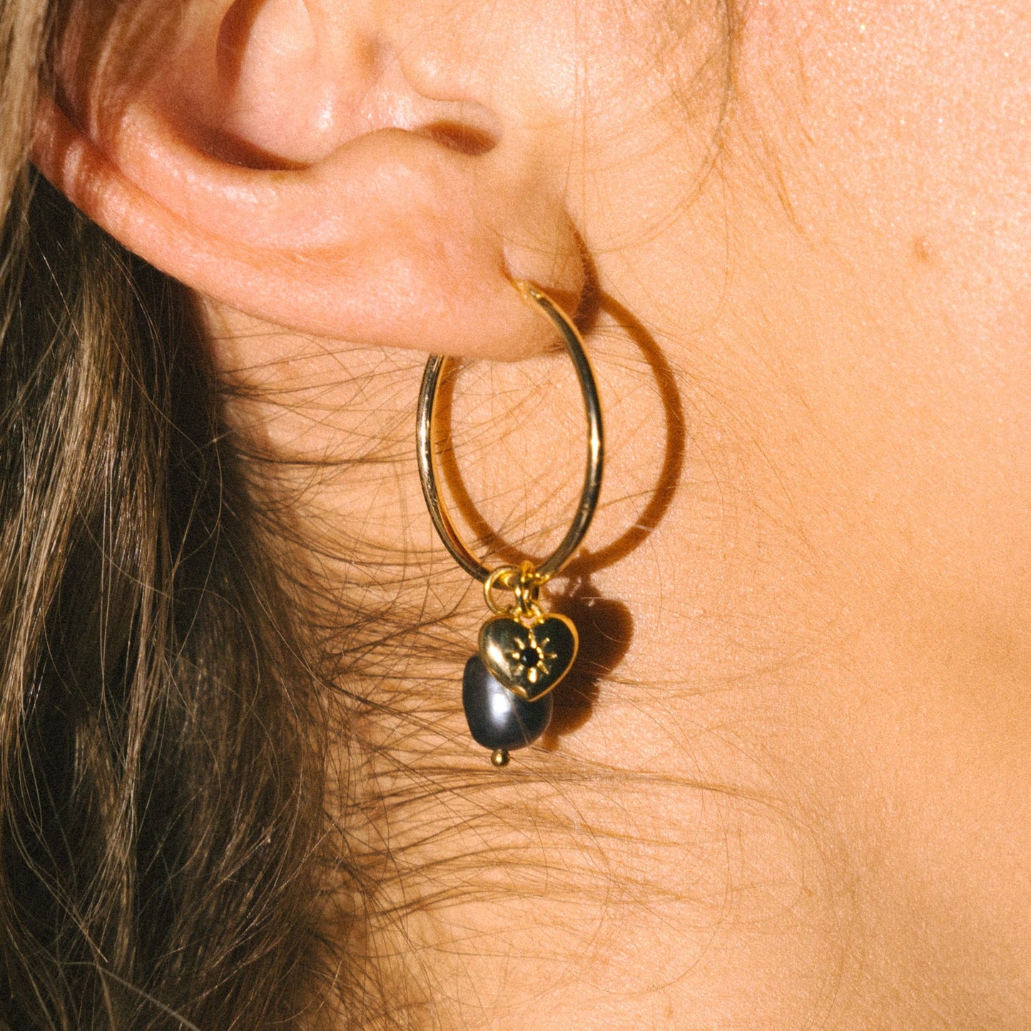 Close-up of a gold hoop earring with a heart-shaped pendant worn by a person.