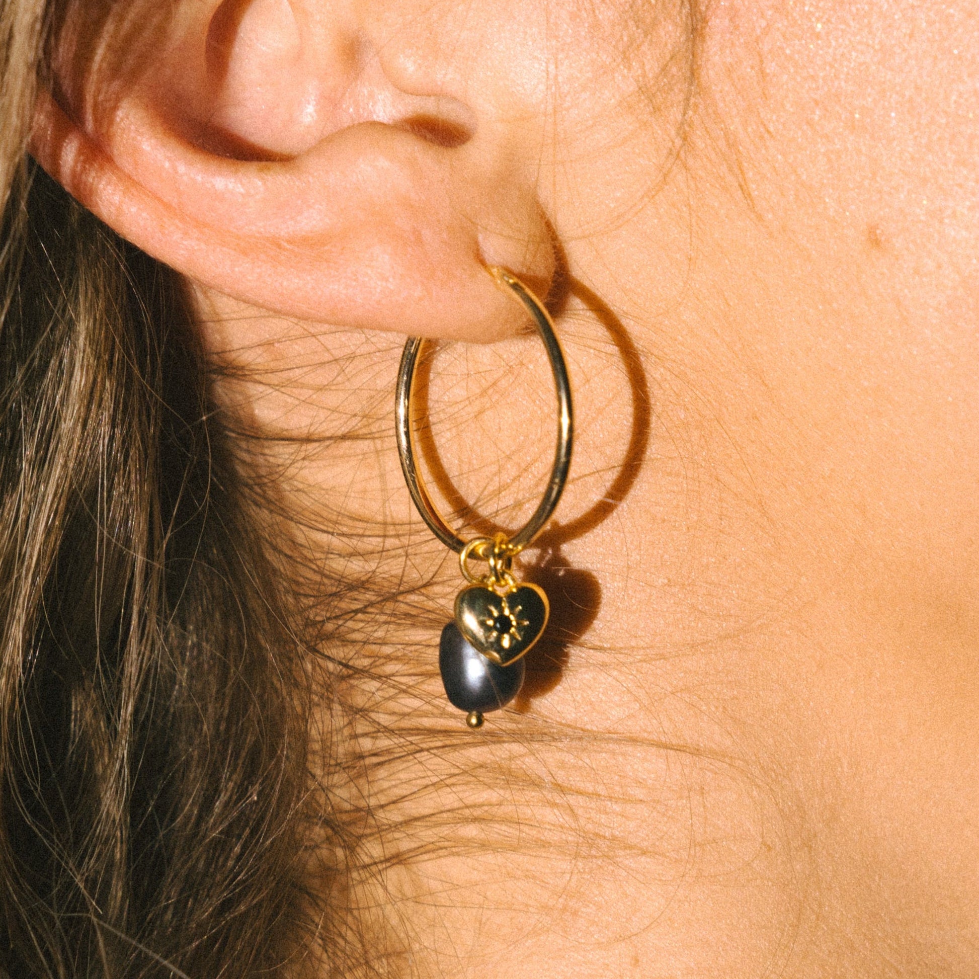 Close-up of a gold hoop earring with a heart-shaped pendant worn by a person.