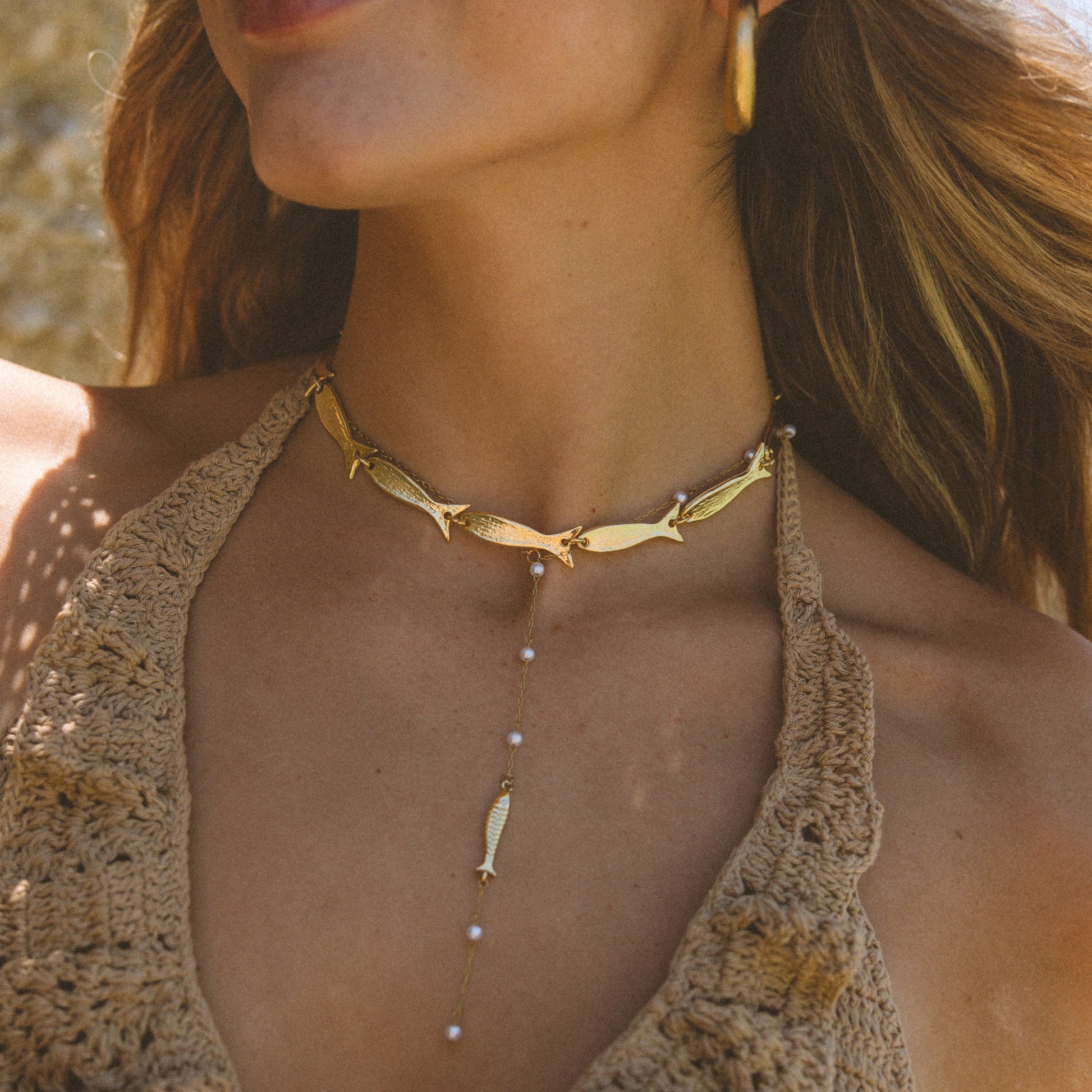 A close-up lifestyle shot of a woman wearing a gold-toned multi-layer necklace. The necklace features a fish-shaped choker, a central drop chain with small pearl-like beads, and a dangling feather-shaped charm.