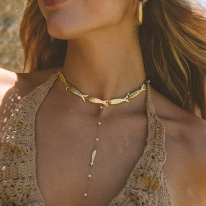 A close-up lifestyle shot of a woman wearing a gold-toned multi-layer necklace. The necklace features a fish-shaped choker, a central drop chain with small pearl-like beads, and a dangling feather-shaped charm.