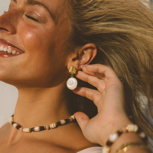 A close-up lifestyle shot of a woman wearing a gold and white spiral shell drop earring, along with a beaded choker and bracelets.