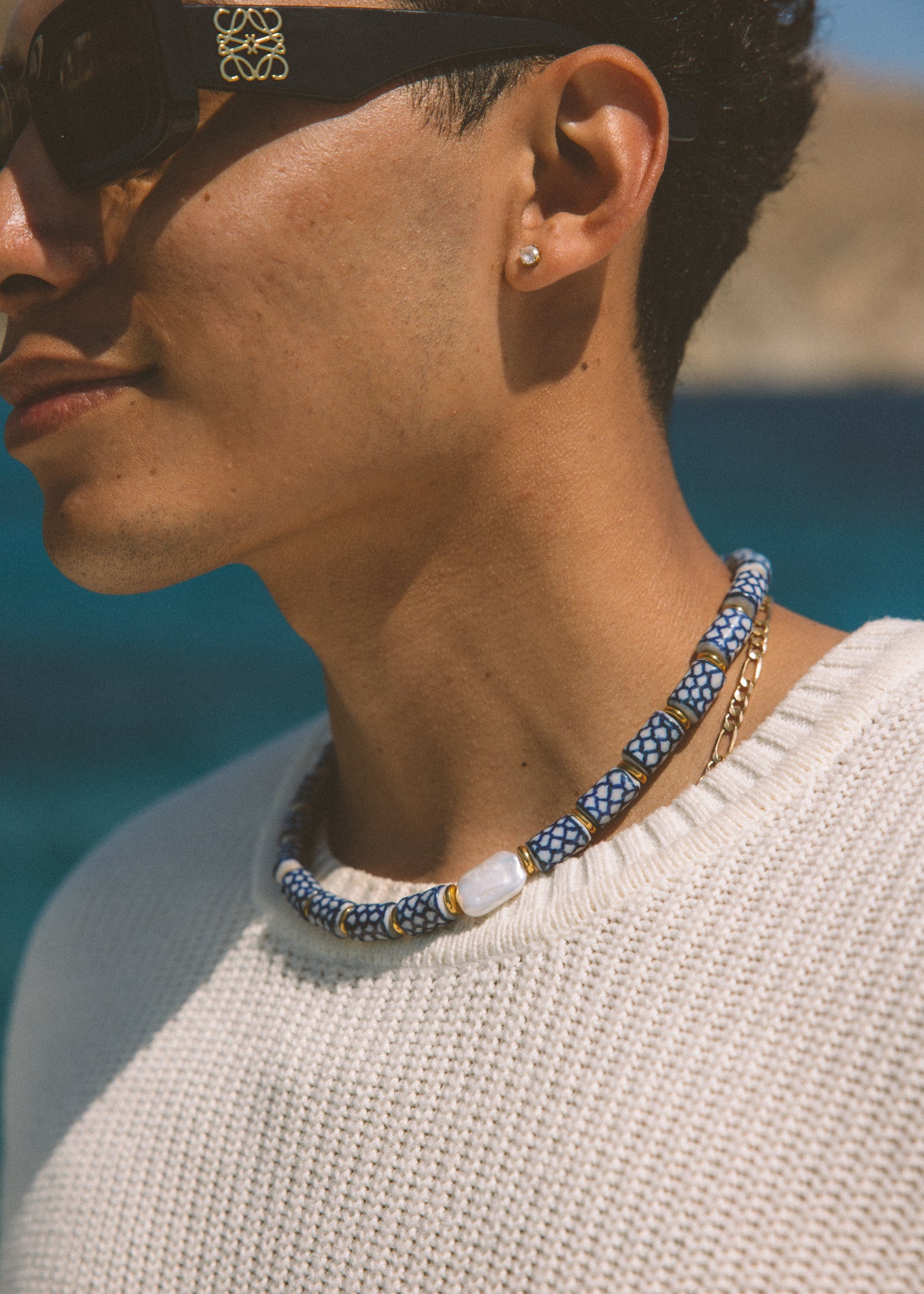 A close-up lifestyle shot of a man wearing a beaded necklace with blue, white, and gold beads, a white knit sweater, and sunglasses. The necklace features a pattern of blue and white beads with a central white stone and gold accents.