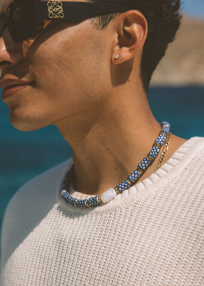 A close-up lifestyle shot of a man wearing a beaded necklace with blue, white, and gold beads, a white knit sweater, and sunglasses. The necklace features a pattern of blue and white beads with a central white stone and gold accents.