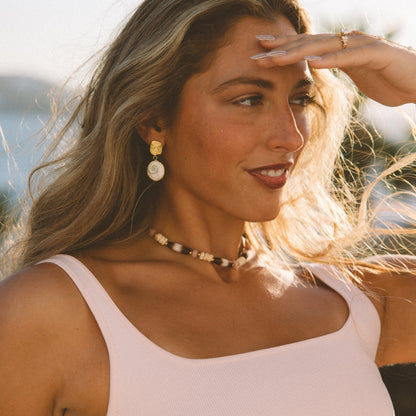 A woman outdoors wearing a pale pink sleeveless top with black fringe trim at the hem, along with a beaded necklace and gold earrings. She is shielding her eyes from the sun with one hand.