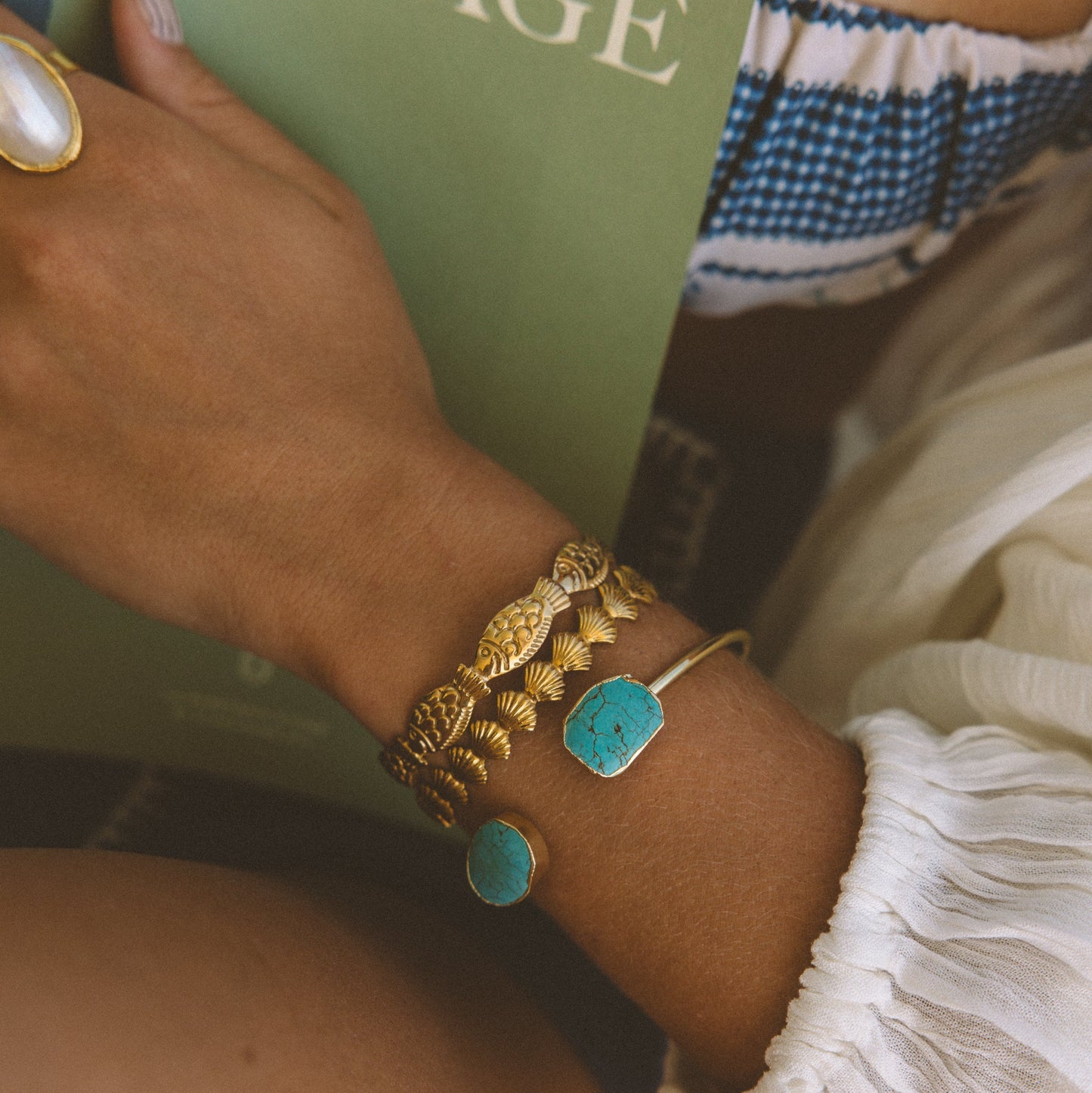 A close-up of a person's wrist wearing a stack of gold bracelets, including one with a turquoise stone and another with a textured gold leaf design, while holding a green book with the word 'VOYAGE' on the cover.