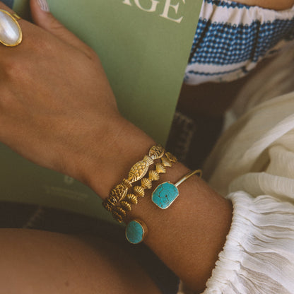 A close-up of a person's wrist wearing a stack of gold bracelets, including one with a turquoise stone and another with a textured gold leaf design, while holding a green book with the word 'VOYAGE' on the cover.