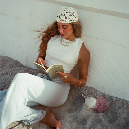 A woman sits on sand against a white wall, reading a book. She is wearing a white sleeveless top and white pants, with a white crocheted head covering and layered necklaces.