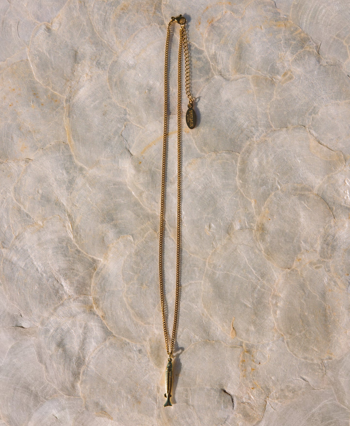 A gold-toned chain necklace with a small fish-shaped pendant and an adjustable chain extension, laid flat on a textured light beige stone surface.