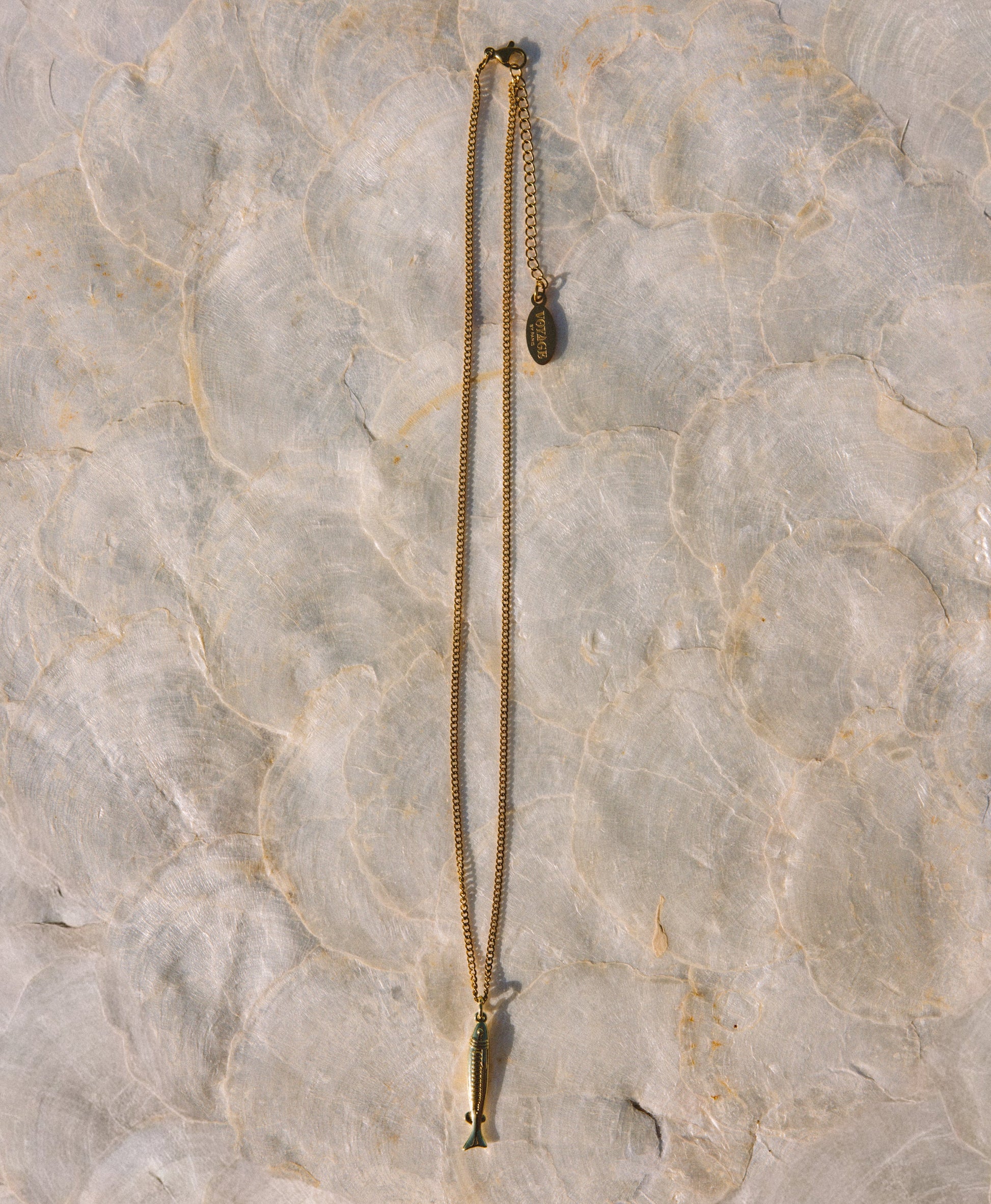 A gold-toned chain necklace with a small fish-shaped pendant and an adjustable chain extension, laid flat on a textured light beige stone surface.