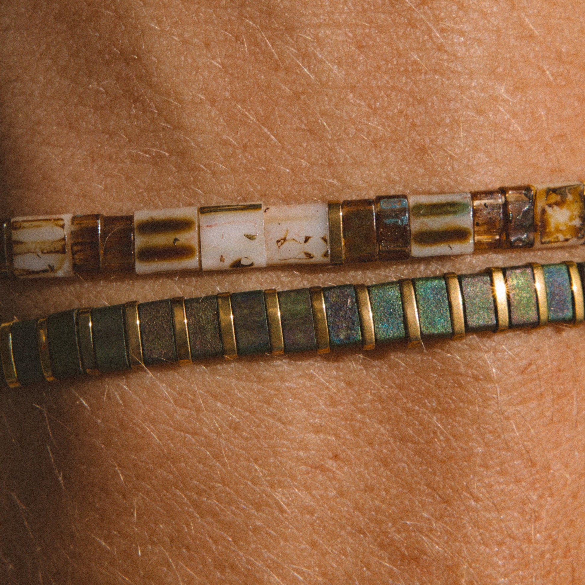 Two beaded bracelets worn on a person's wrist, featuring rectangular beads in earthy brown, cream, and green with gold-toned borders.