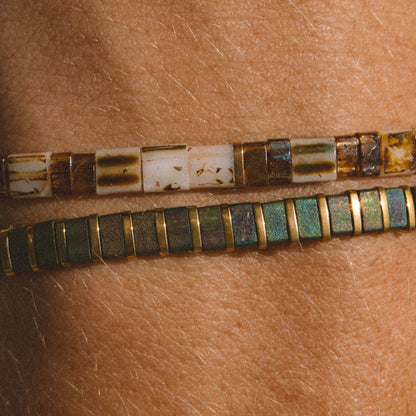 Two beaded bracelets worn on a person's wrist, featuring rectangular beads in earthy brown, cream, and green with gold-toned borders.