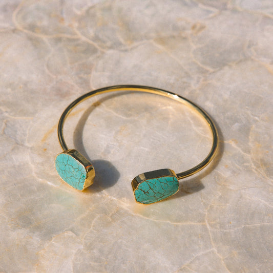 A gold open cuff bracelet with two flat, hexagonal turquoise-colored stone end caps, resting on a light beige marble surface.