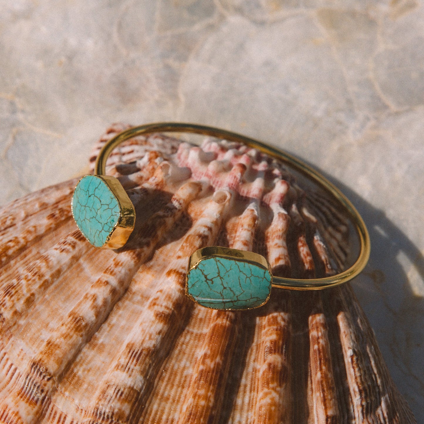 Gold cuff bracelet with two oval turquoise stone settings, displayed on a large seashell against a light stone surface.
