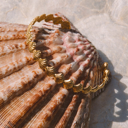 A gold-toned bangle bracelet with a textured, ridged shell-like design, displayed on a large brown seashell against a light stone surface.