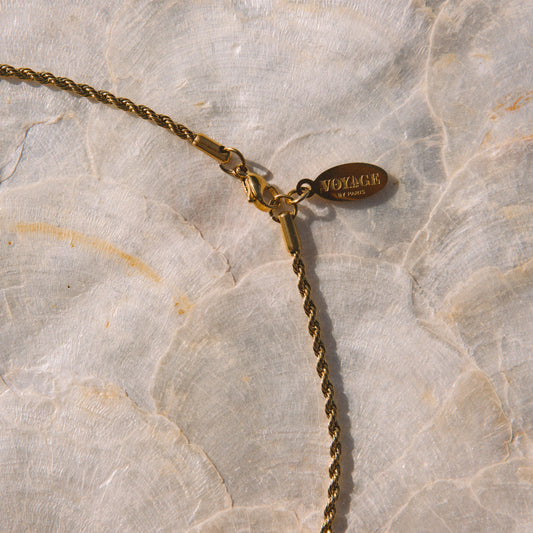 Close-up of a gold chain necklace with a lobster clasp and a small oval brand tag, laid on a textured off-white surface.