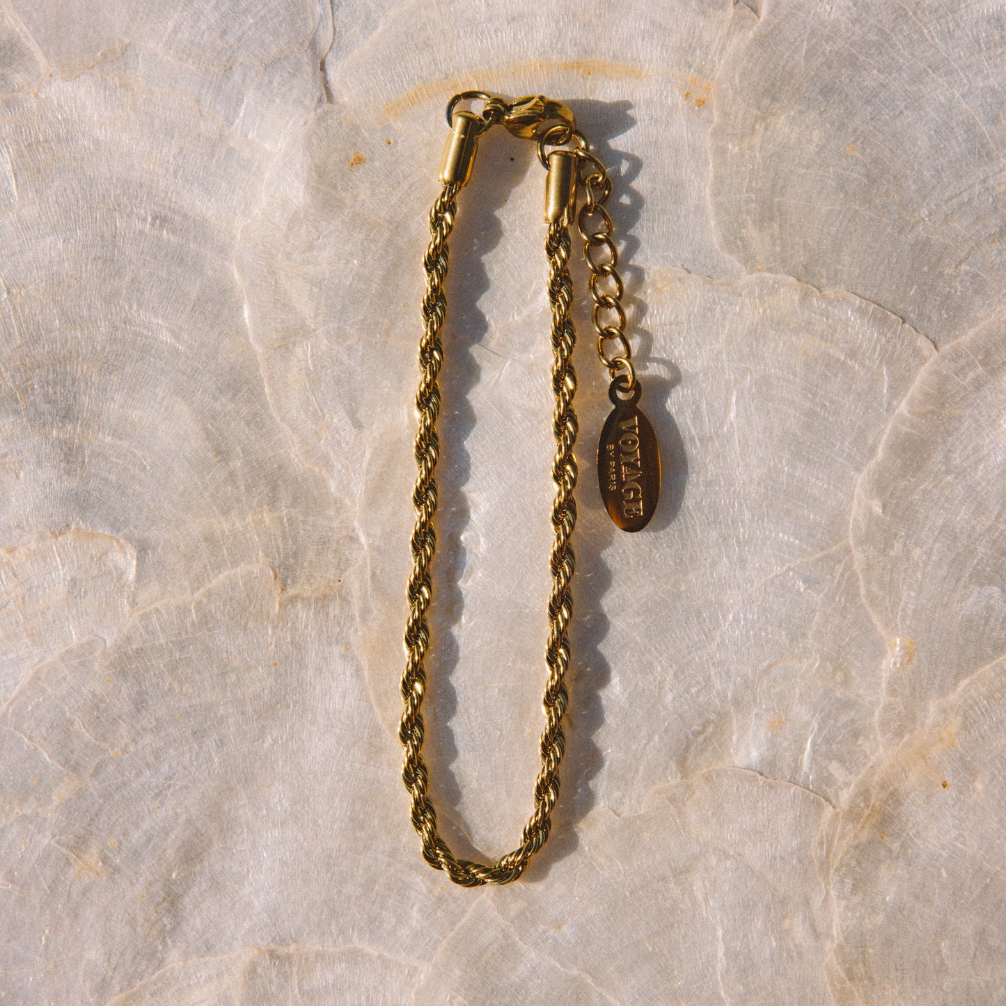 A gold-toned chain bracelet with a lobster clasp and an adjustable extender chain featuring a small oval charm, displayed on a light beige stone surface.