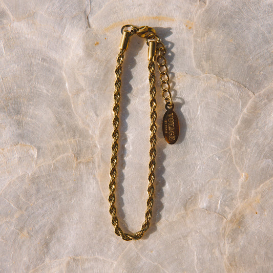 A gold-toned chain bracelet with a lobster clasp and an adjustable extender chain featuring a small oval charm, displayed on a light beige stone surface.