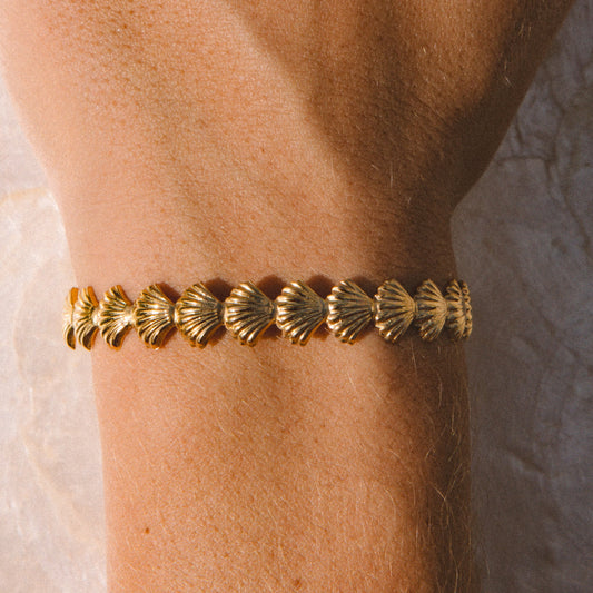 A gold bracelet with a row of small shell motifs, shown worn on a person's wrist against a neutral stone-textured background.