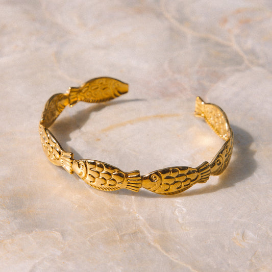A gold-toned open cuff bracelet with embossed fish motifs, displayed on a light beige marble surface.