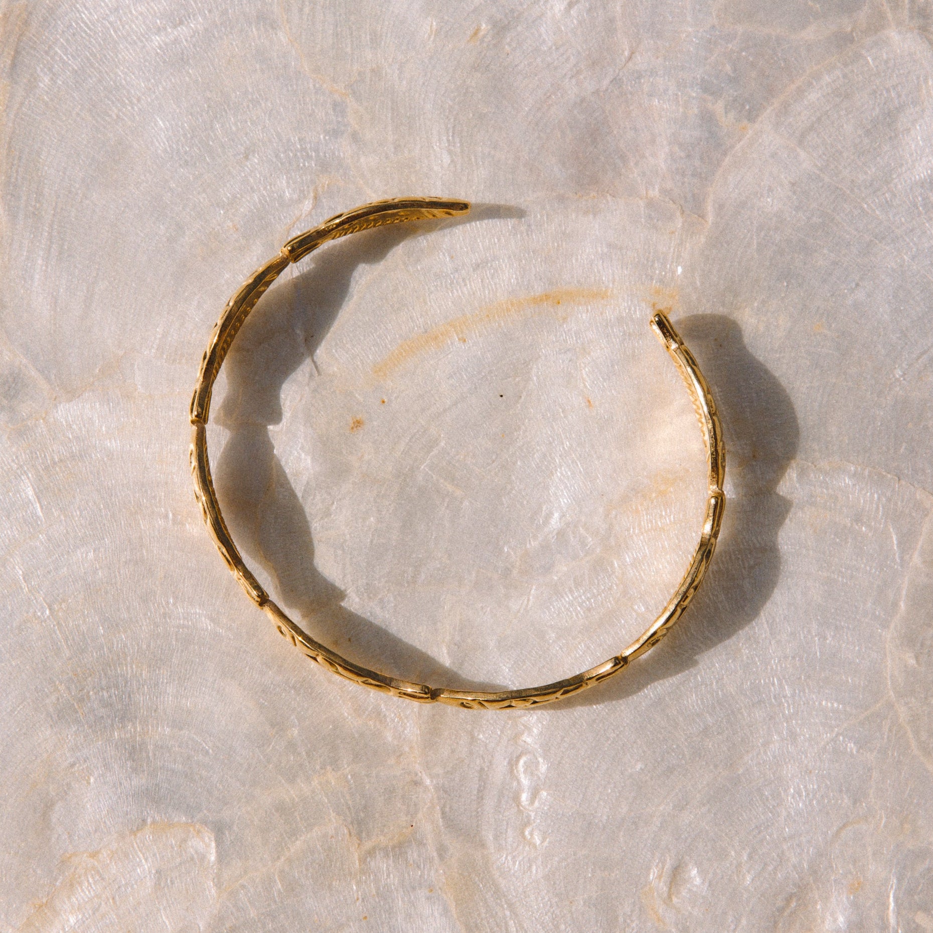 A single thin gold bangle bracelet with a textured, hammered finish, displayed on a light beige marble surface.