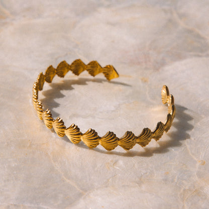 A gold-toned open cuff bracelet with a textured, ridged shell-like pattern, displayed on a light beige stone surface.