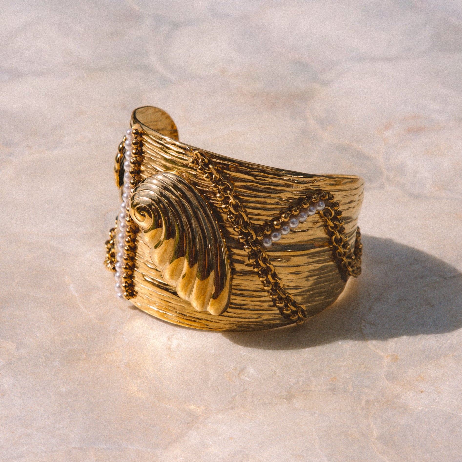 A wide gold cuff ring with textured horizontal grooves, photographed on a light stone surface.