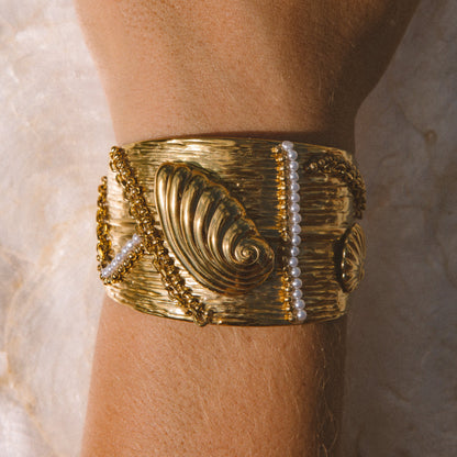 A wide gold cuff bracelet with textured spiral motif and bead details, worn on a person's wrist against a light background.