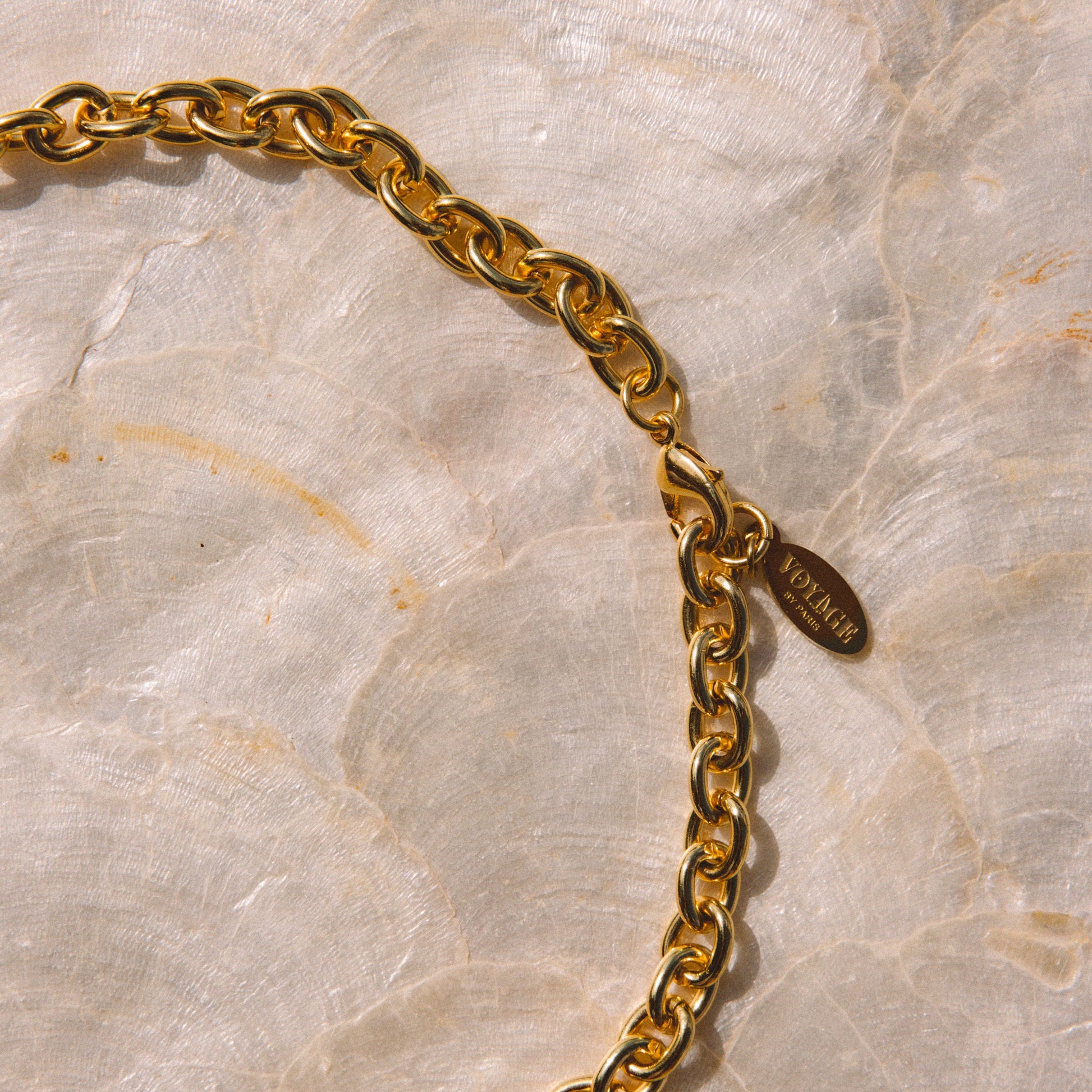 A close-up of a gold-tone chain necklace with a small oval brand tag, laid on a light beige marbled surface. The tag is engraved with the brand name Versace.