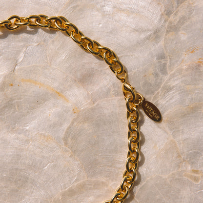 A close-up of a gold-tone chain necklace with a small oval brand tag, laid on a light beige marbled surface. The tag is engraved with the brand name Versace.