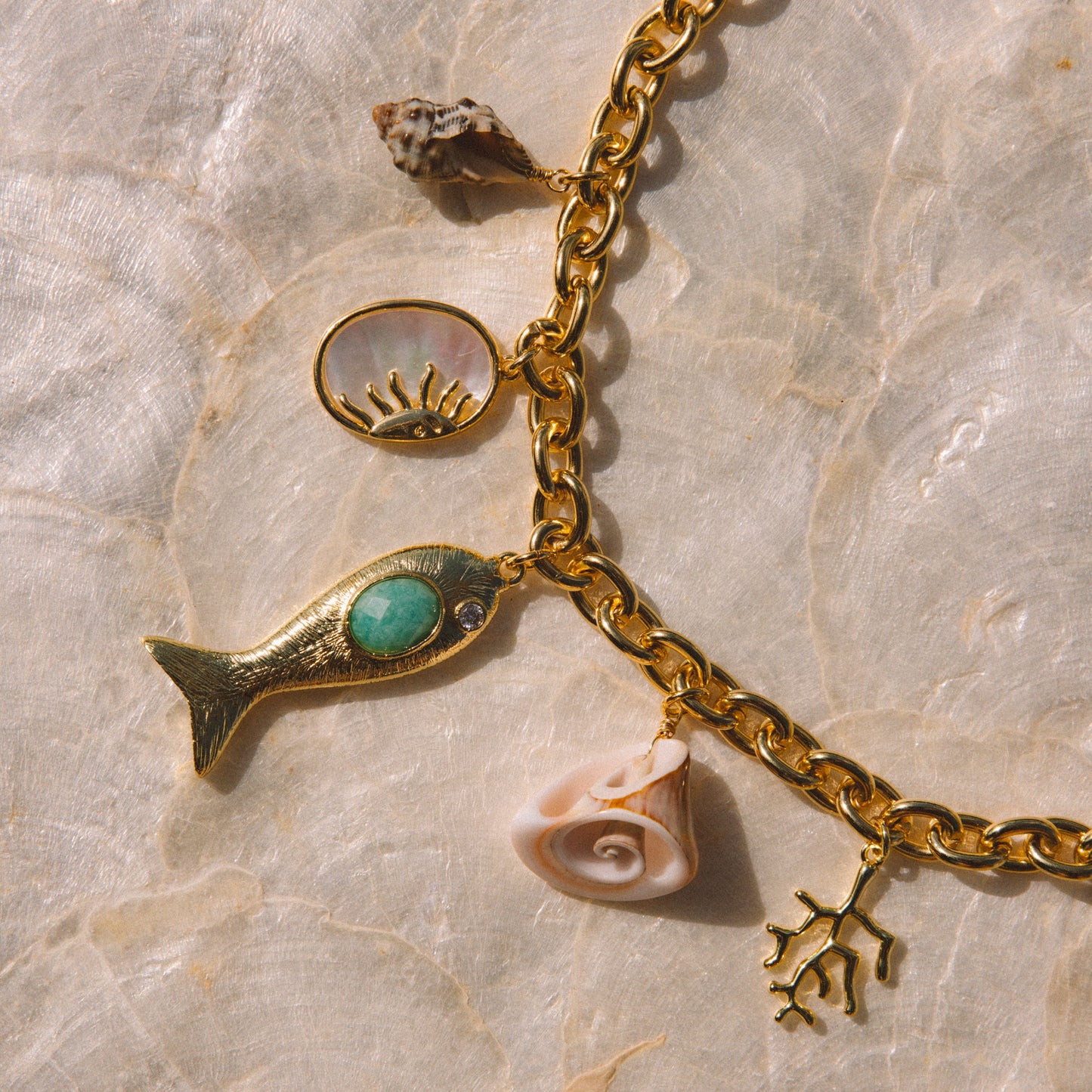 Gold-tone chain necklace with seashell and sea-themed charms, including a fish with a turquoise stone, a shell charm, a spiral shell charm, and a coral-like charm, displayed on a textured beige surface.